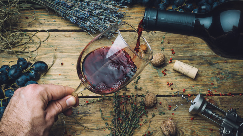 An individual pouring red wine for tasting in front of grapes, herbs, and corkscrew