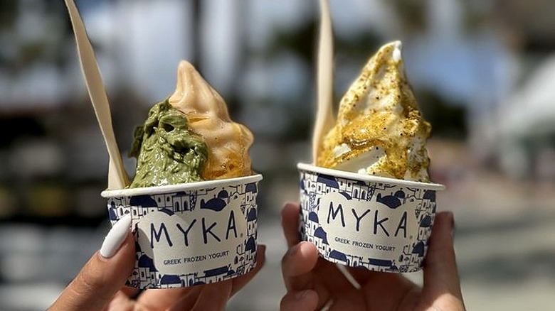 Two hands holding up small containers of Myka yogurt with toppings.