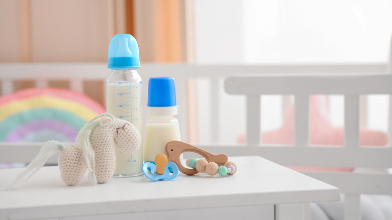 Bottles of baby milk formula, toys, and a pacifier on a nightstand by a crib
