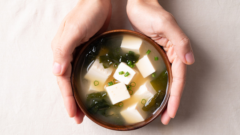 A person's hands holding a cup of miso soup with tofu and spring onions