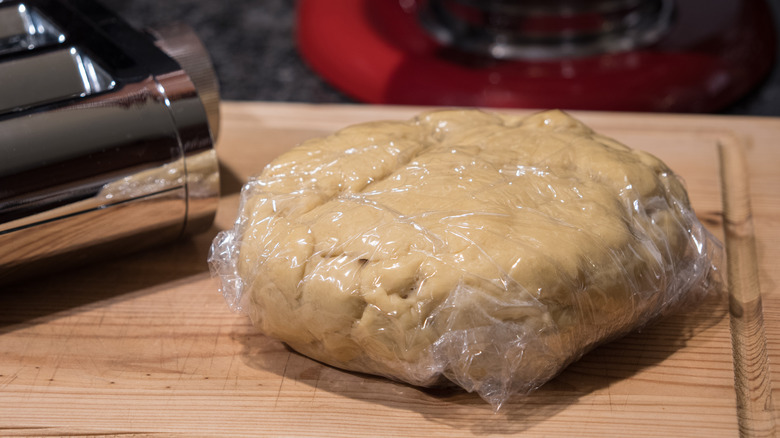 Pasta dough wrapped in plastic wrap resting on a wooden cutting board