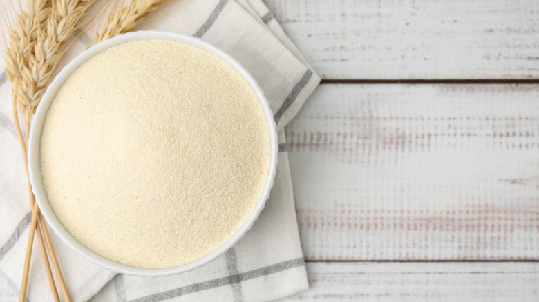 A bowl of semolina flour next to a sprig of wheat and a dish towel