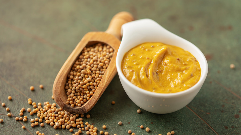 Mustard in a white bowl and a wooden scoop full of mustard seeds