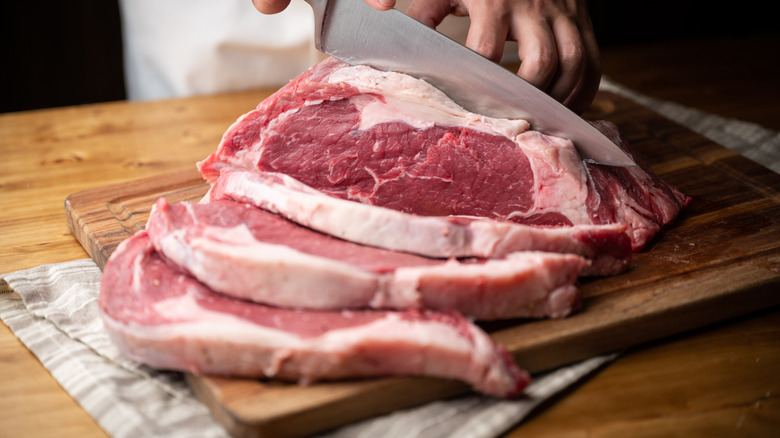 Stainless steel knife slicing a whole ribeye into steaks on a cutting board