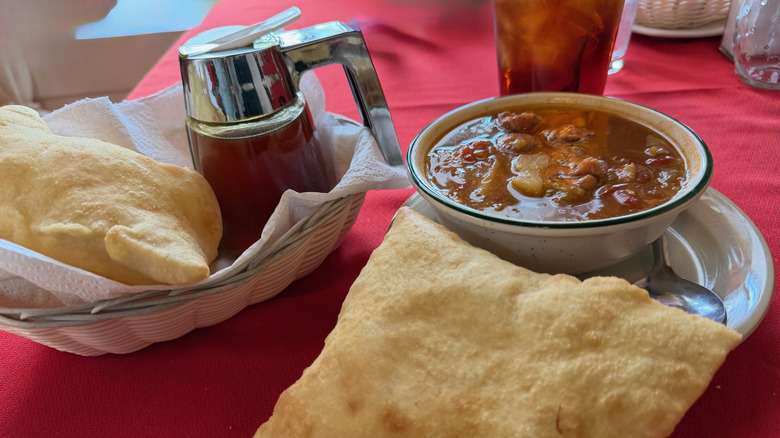 two golden sopaipillas, a container of honey, and a bowl of green chile stew on a red tablecloth