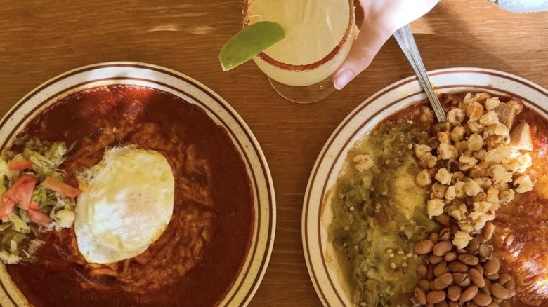 Red chile enchiladas topped with egg on a white plate and green chile, pinto beans, and nixtamal corn on a separate white plate