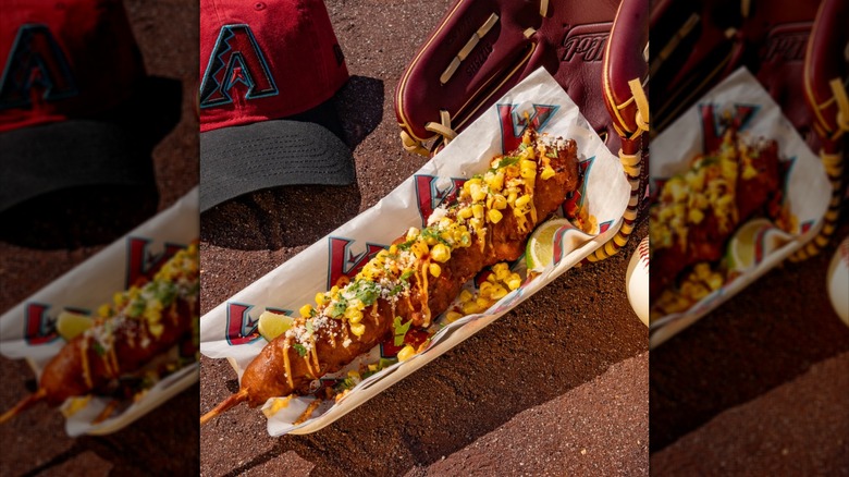 Footlong elote corndog in paper serving tray next to baseballs, hat, and glove.