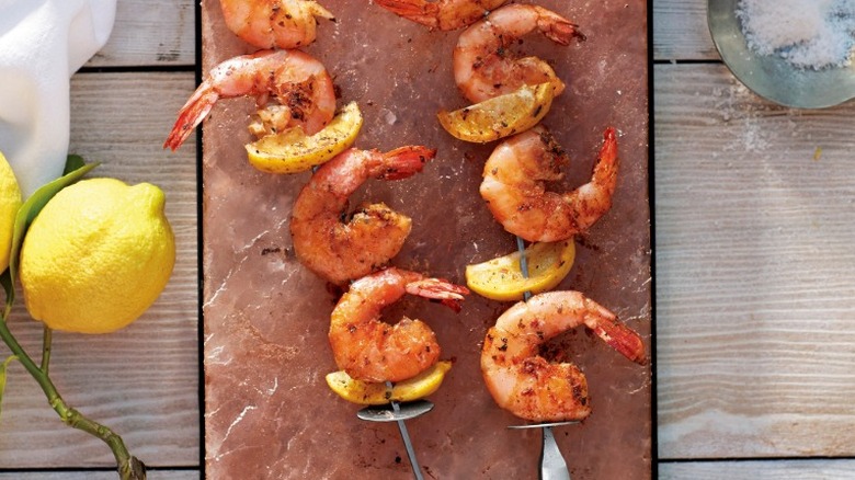 Kebabs of shrimp and citrus laying on Himalayan pink salt plank with whole lemon nearby.