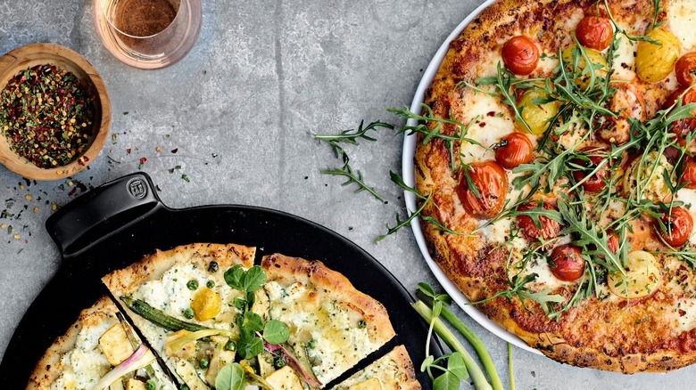 Two artisanal pizzas with veggies and herbs and a bowl of spices nearby.