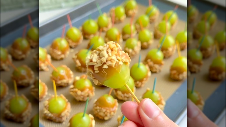 Person holding a caramel and peanut coated green grape in front of a tray of caramel grapes