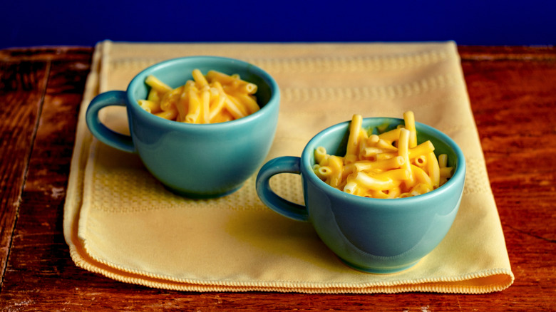 two teal mugs of mac and cheese on yellow cloth on wooden table