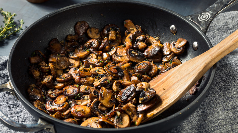 A non-stick skillet full of sauteed, browned mushrooms with thyme on a denim cloth, over a grey table, thyme sprigs in the background.
