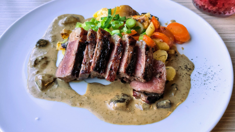 A full plated meal of medium rare sliced steak surrounded by roasted vegetables and a creamy mushrooms sauce.