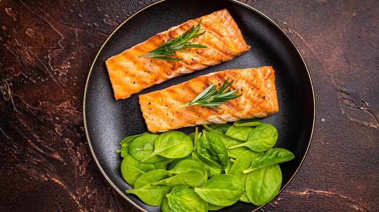 A plate of two pieces of grilled salmon with beautiful caramelization, topped with sprigs of rosemary, leaves of baby spinach below on a brown stone table.