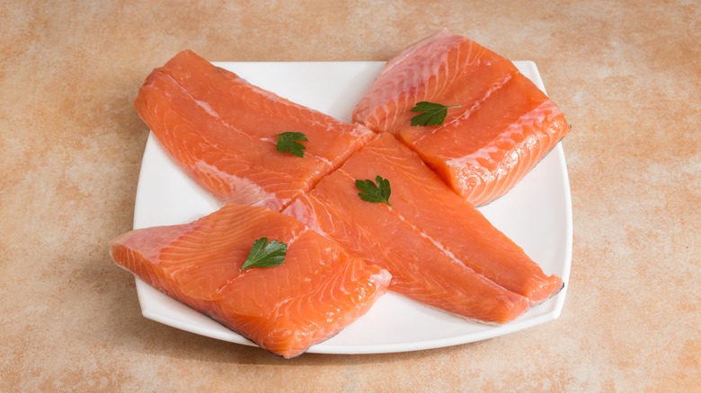 Four cuts of bright pink, raw salmon loin on a white plate, topped with parsley leaves.