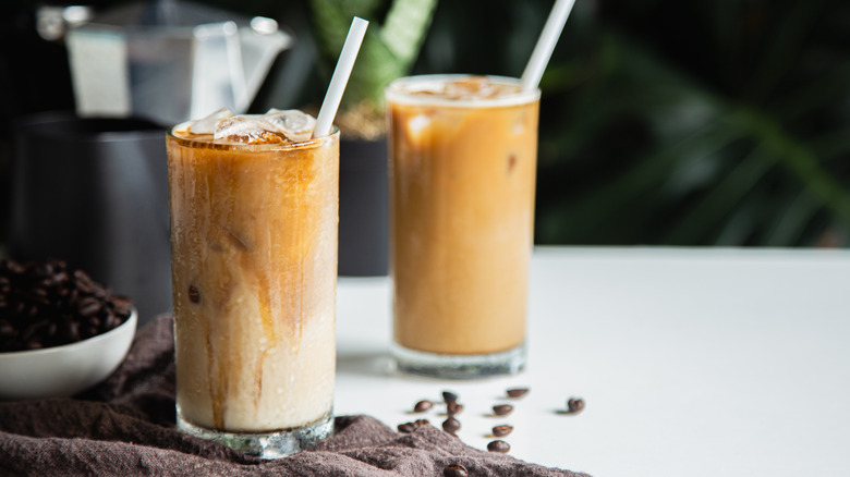 Two glasses of iced lattes with straws on counter with loose coffee beans.