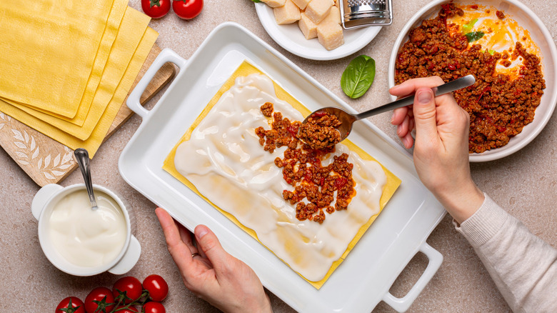 Hands assembling homemade lasagne with layers of béchamel and ragù.