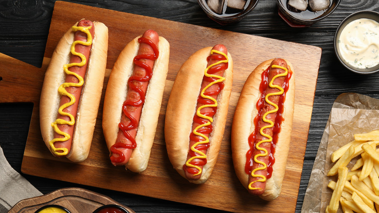 A wooden cutting board with four hot dogs dressed with mustard and ketchup