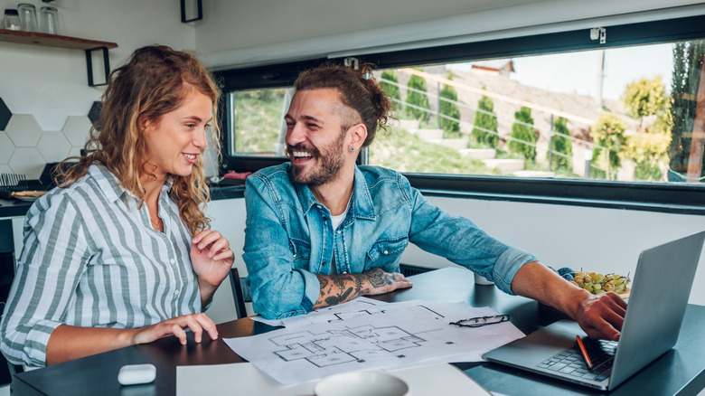 Couple sitting at a table looking at blueprints, smiling, while man points to open laptop.
