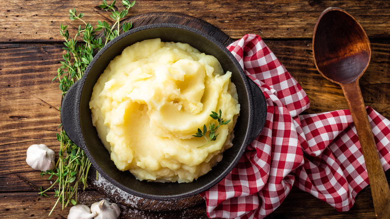 A bowl of fluffy mashed potatoes on a wooden table in a rustic setting, with a spoon, cloth, herbs, and garlic on the side