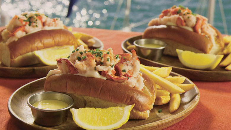 Three lobster rolls on plates in front of a body of water
