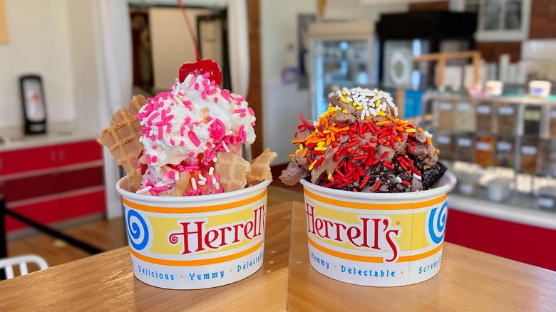 Two Herrell's branded cups of ice cream with toppings sitting on a wooden surface