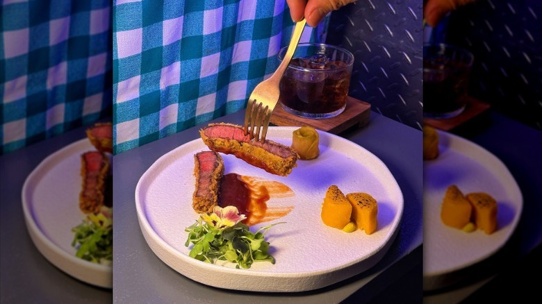 Fork picking up a slice of grilled wagyu from a white plate with microgreens and other garnishes