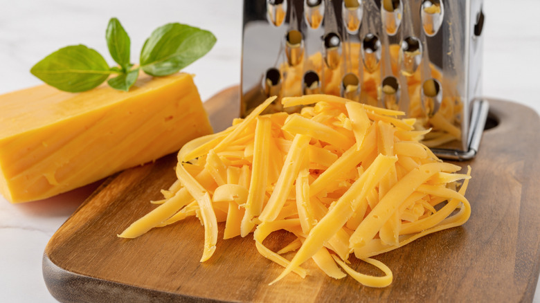 A brick of cheddar sitting next to a grater with grated cheddar in the foreground.