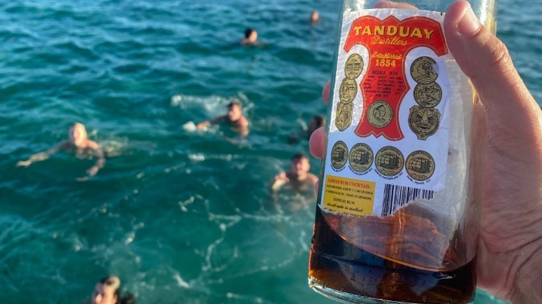 A person holding a bottle of Tanduay rum over the side of a boat as people swim in the water behind it.