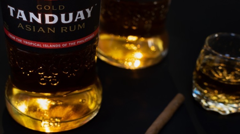 A bottle of Tanduay gold rum sitting next to a fresh pour on a dark surface.