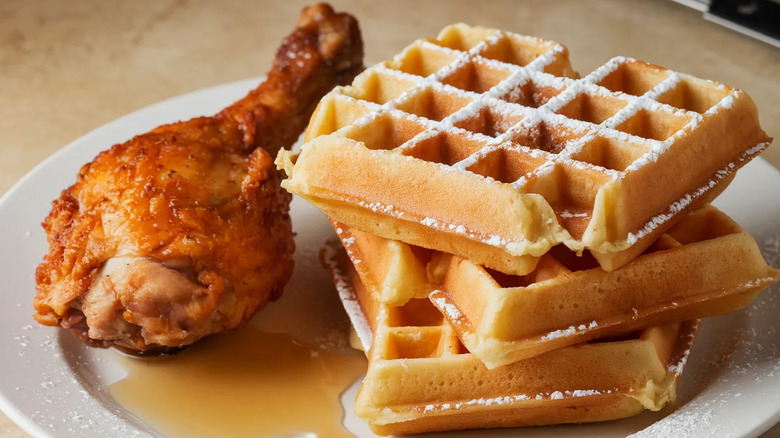 A fried chicken drumstick next to a stack of square waffles with syrup on the bottom of the plate