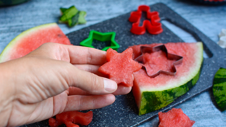 hand holding a star shaped watermelon piece, with watermelon wedges and cookie cutters in the background over a chopping board