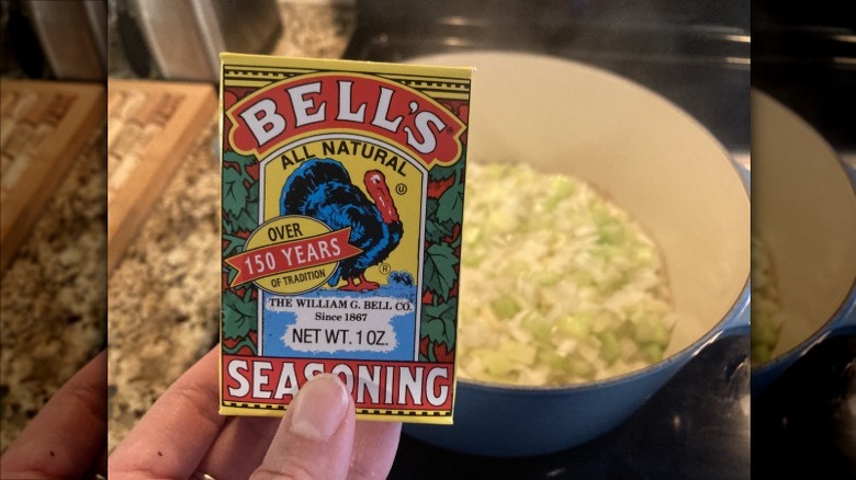Close-up of a hand holding a box of Bell's Seasoning in front of a pot filled with celery and onions