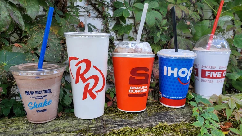 5 fast food chain milkshakes lined up next to each other in an outdoor setting.