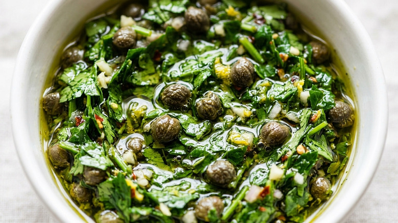 Close-up of Italian salsa verde with capers, lemon peel, and fresh green herbs like parsely in a white ceramic ramekin