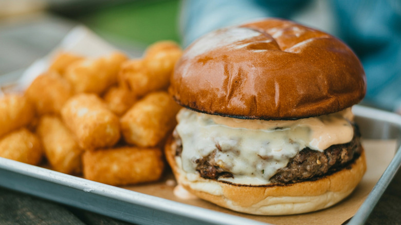A cheeseburger with a toasted bun next to tater tots on a metal tray