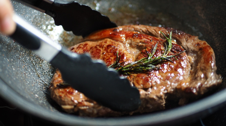 Searing a a ribeye in a hot skillet, covered in rosemary.
