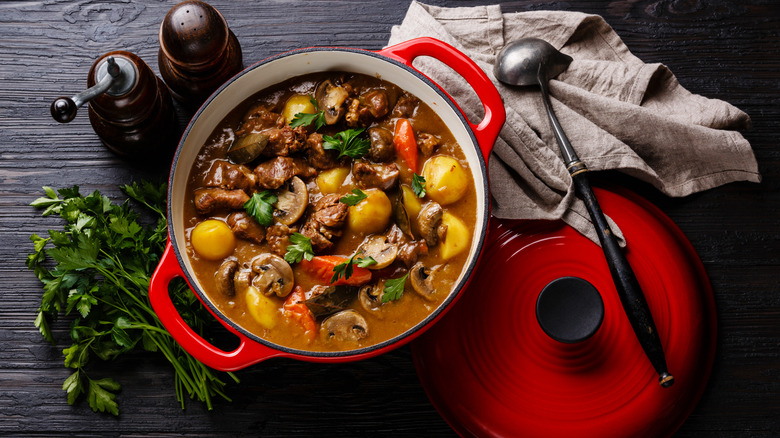 Red Dutch oven pot of beef stew on a dark gray wooden table with herbs, salt and pepper shakers, and a ladle on a cloth napkin