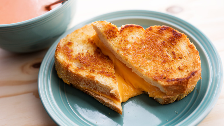 A sliced, oozing grilled cheese sandwich on a plate with a bowl of soup in the background