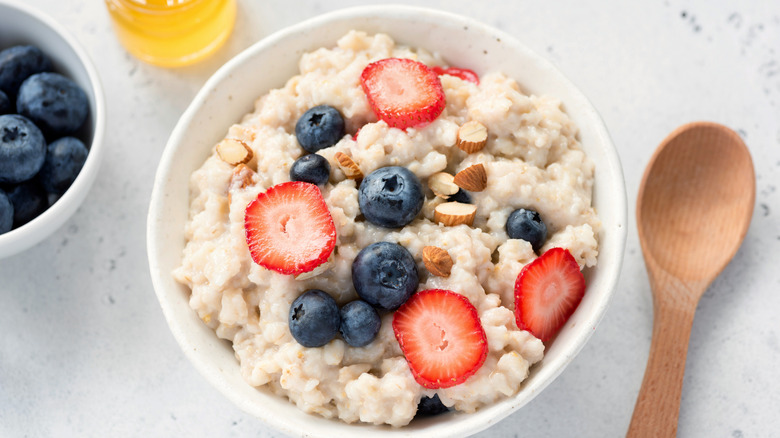 A bowl of oatmeal on a white countertop, topped with sliced strawberries, sliced almonds, and whole blueberries, surrounded by a wooden spoon, a jar of honey, and a bowl of blueberries.