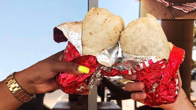 Two people tapping burritos in red wrappers together