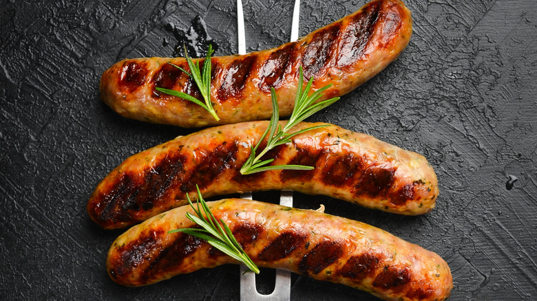 Three perfectly charred breakfast sausages, pierced by a two-prong fork, set against a black textured background