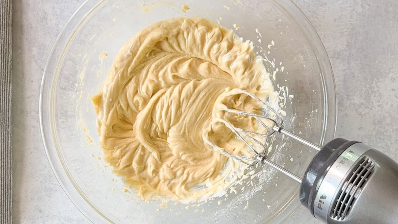 Sour cream mixed into blueberry crumb cake batter in glass bowl with hand mixer