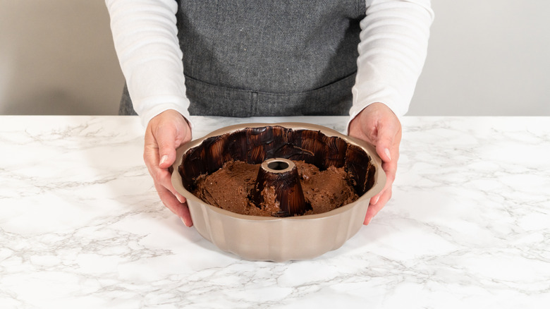 Person holding a bundt pan filled with chocolate cake batter that's resting on a marble countertop