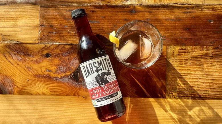 Bottle of Barsmith Hot Honey Old Fashioned lying on wooden tabletop with a cocktail made