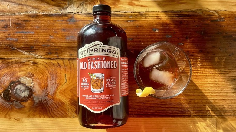 Bottle of Stirrings Simple Old Fashioned Cocktail Mix lying on a wooden tabletop with a cocktail made