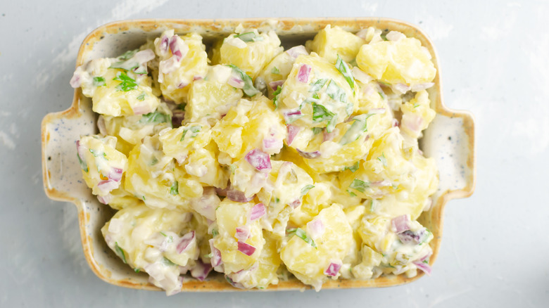 Cold potato salad with purple onion, pickles, and parsley in a white rectangular bowl on a gray table.