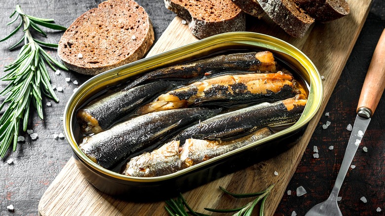 Opened can of sardines next to slices of brown bread on a wooden serving tray