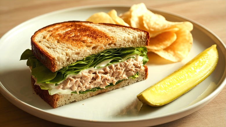 Half of a toasted tuna salad sandwich with potato chips and a pickle spear on a cream-colored plate