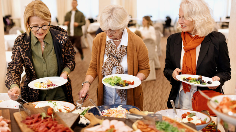 Three older women holding plates in front of a buffet getting food.
